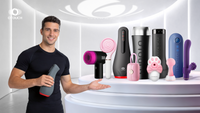A smiling man in a black sports T-shirt holds a gray sex toy in his left hand and extends his right hand to the right. Behind him, on a white display stand, stands a series of OTOUCH brand sex toys. The background is a modern, futuristic white room.