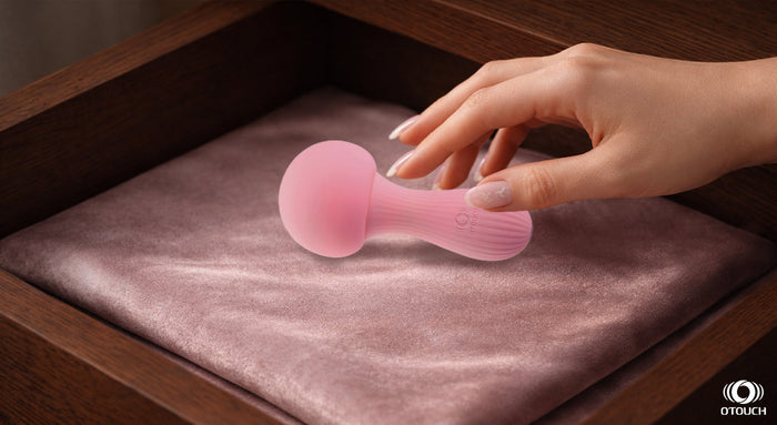 A hand reaching into a wooden nightstand drawer to pick up a pink OTouch wand massager resting on a mauve satin cushion.